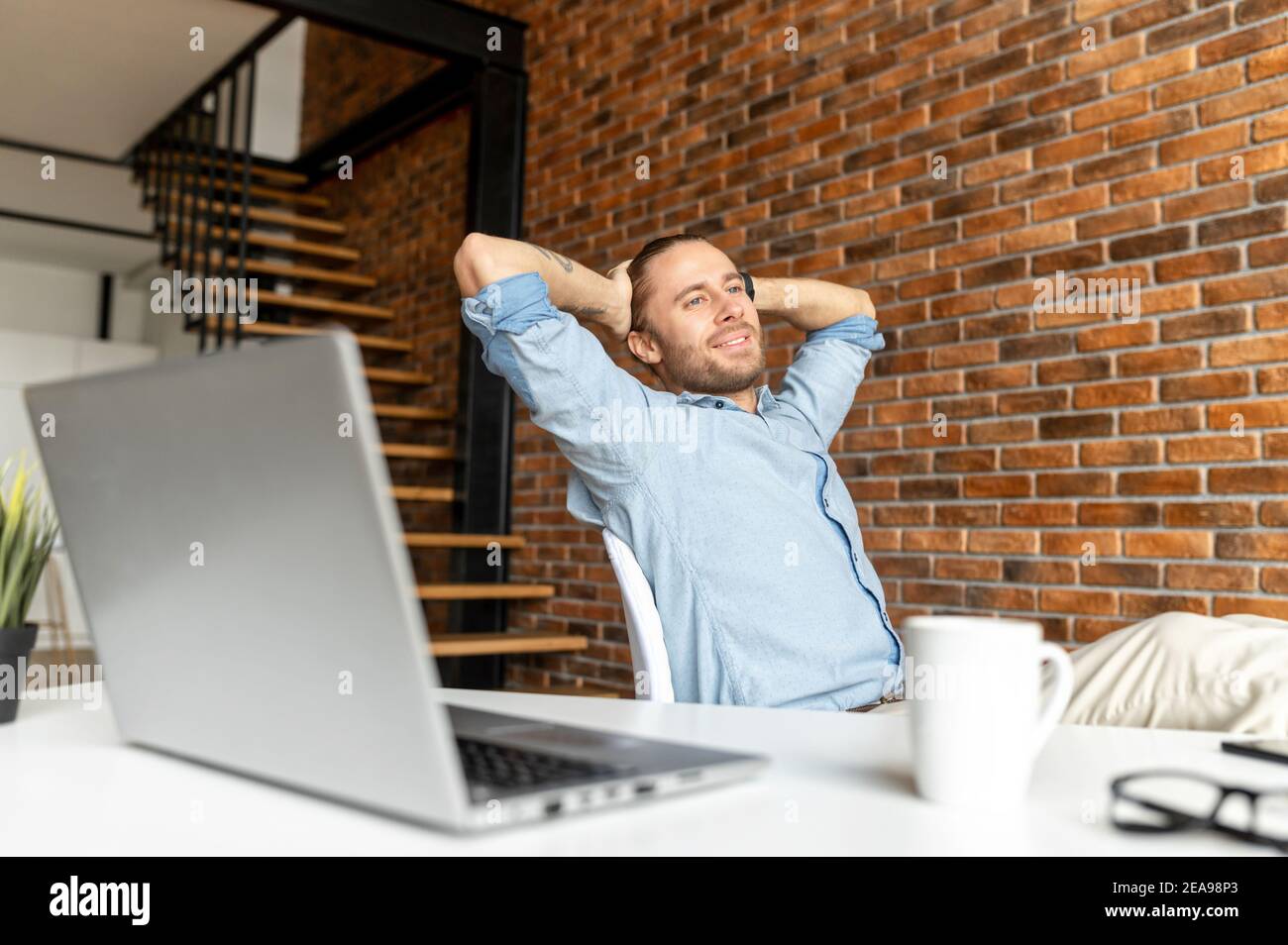 Satisfaction homme d'affaires assis dans une posture confiante avec un pied sur la table et se détend, penché en arrière dans la chaise, tient ses mains derrière sa tête, a Banque D'Images