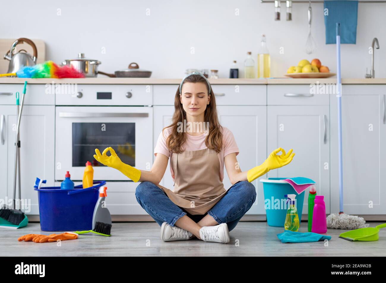 Jeune femme de ménage entourée de fournitures de nettoyage méditant sur le sol à la cuisine, prenant pause des tâches de la maison Banque D'Images