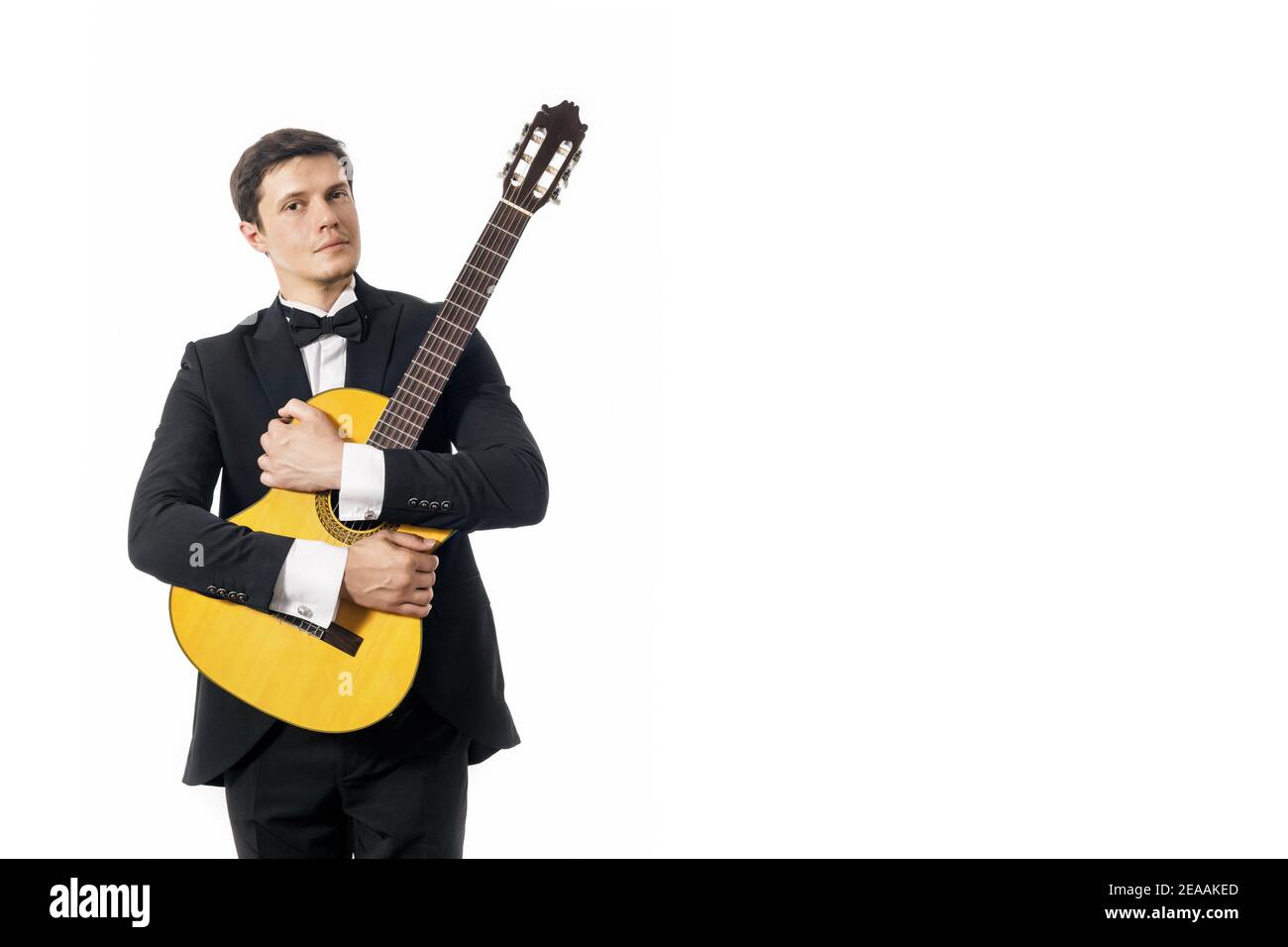 Beau jeune homme en costume classique noir avec noeud papillon posant avec guitare classique en studio sur fond blanc. Banque D'Images