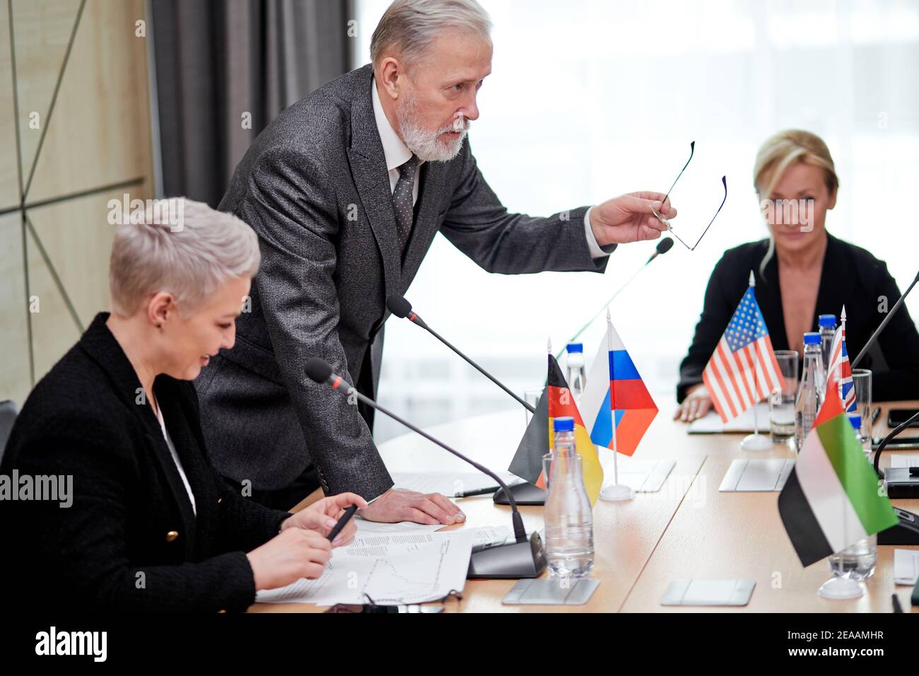 un homme politique senior offre son plan d'action et partage son opinion, un homme âgé en costume s'adressant à un groupe multiethnique de partenaires assis à son bureau en conseil d'administration Banque D'Images