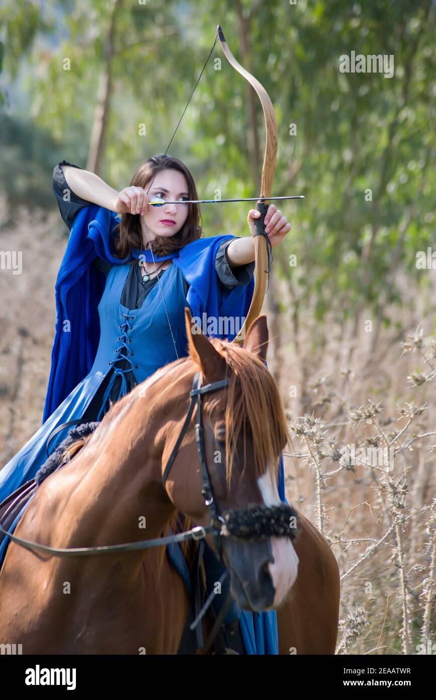 Archer girl en robe bleue de style médiéval, visez à tirer avec une flèche et un arc tout en montant un cheval Banque D'Images