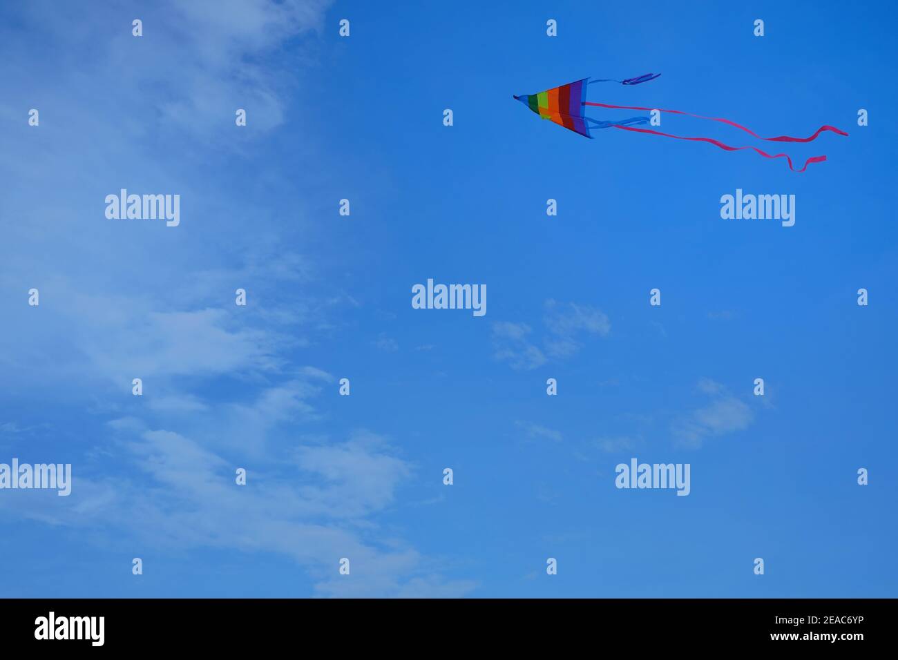 Un cerf-volant en forme de triangle coloré avec un motif arc-en-ciel et une longue queue vole contre un ciel bleu clair et des nuages blancs moelleux. Banque D'Images