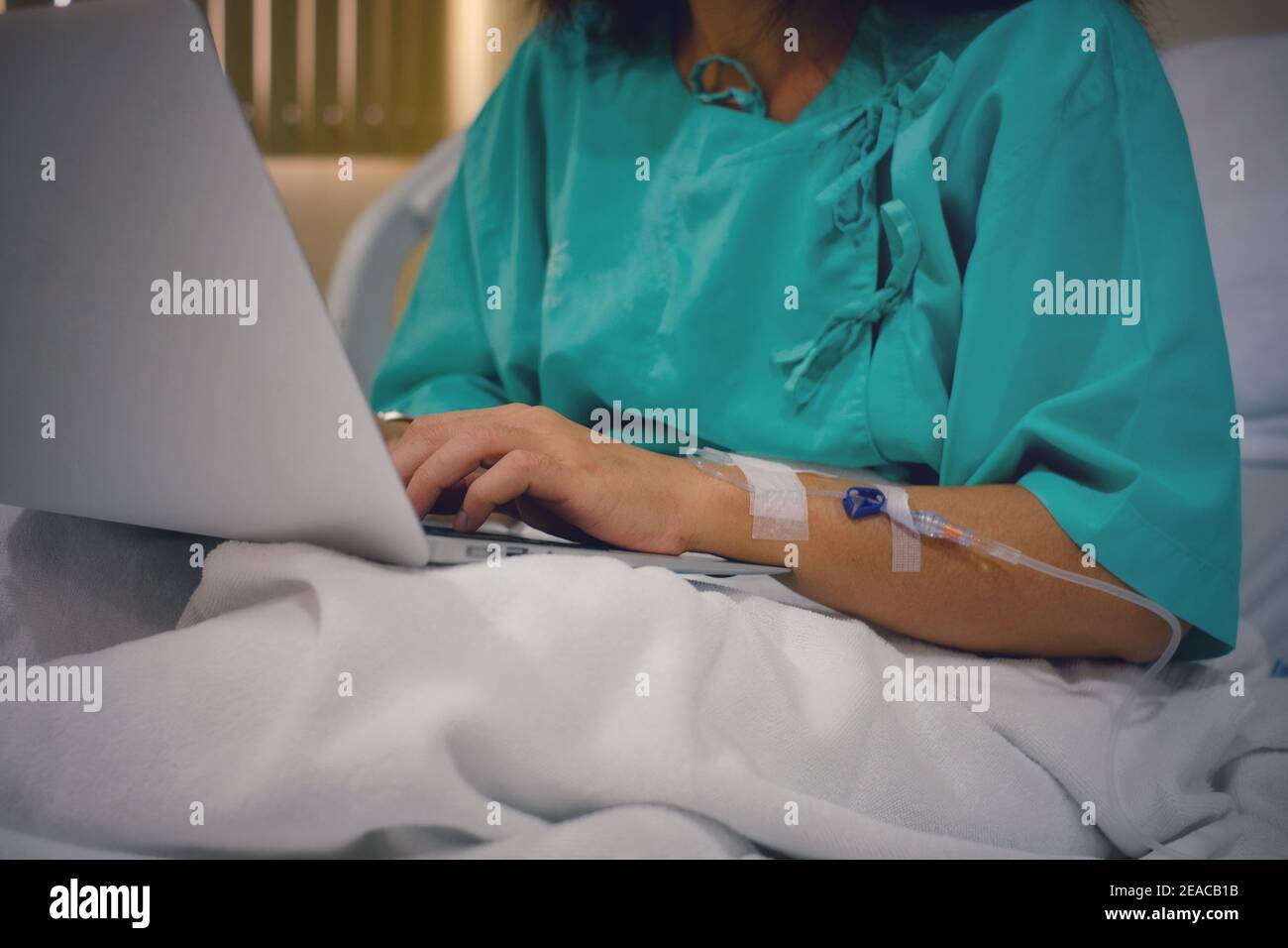 Un demi-coup de corps d'une femme d'affaires de travail assise sur son lit d'hôpital avec injection IV dans son bras, se rétablissant de la chirurgie tout en utilisant son lapte Banque D'Images