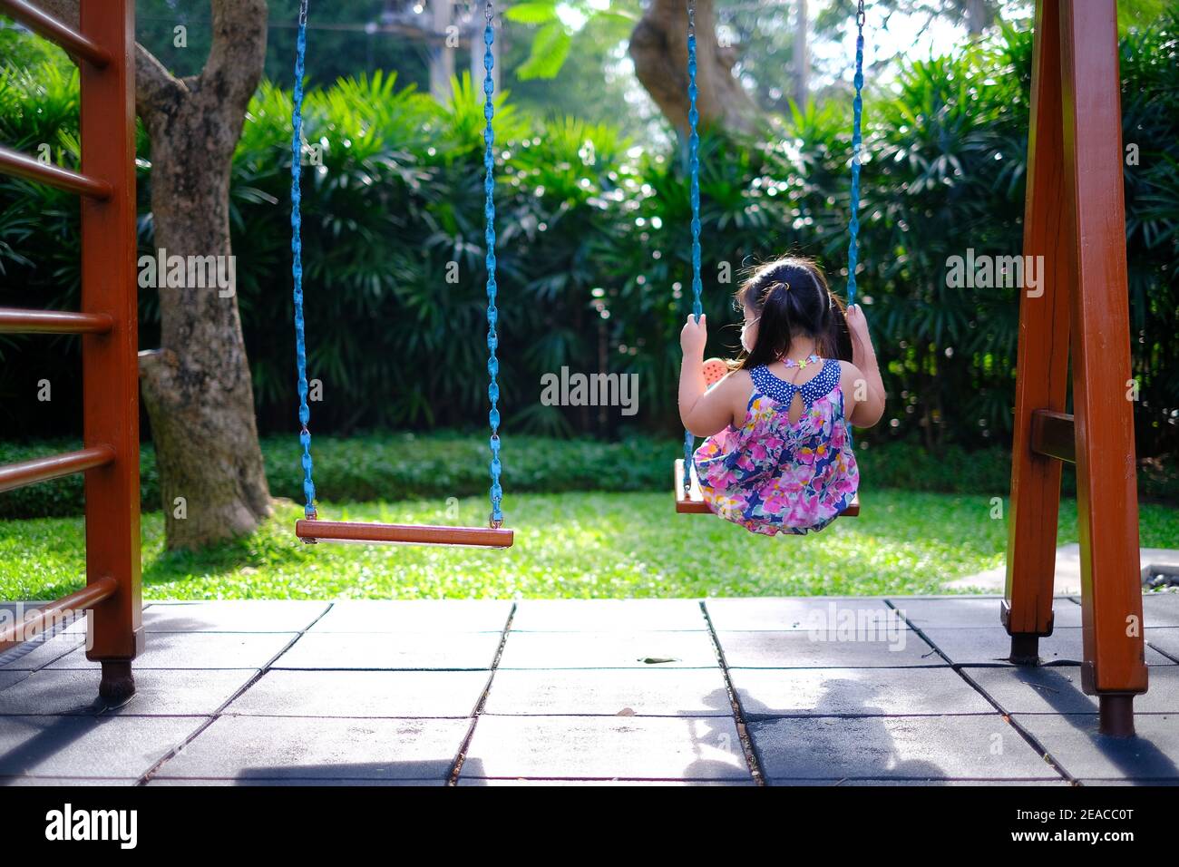 La vue arrière d'une jeune fille asiatique solitaire jouant sur une balançoire par elle-même dans une aire de jeux vide par une journée ensoleillée. Banque D'Images