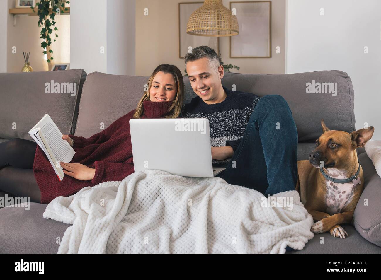 Un couple heureux et leur chien se détendent sur le canapé, lisant un livre et utilisant un ordinateur portable, ayant un bon moment ensemble à la maison. Concept de vie de couple Banque D'Images