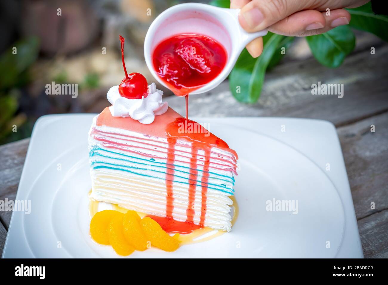 Gâteau à la crêpe arc-en-ciel et une cerise avec sauce aux fraises à la main sur le dessus, fond de table en bois. Banque D'Images