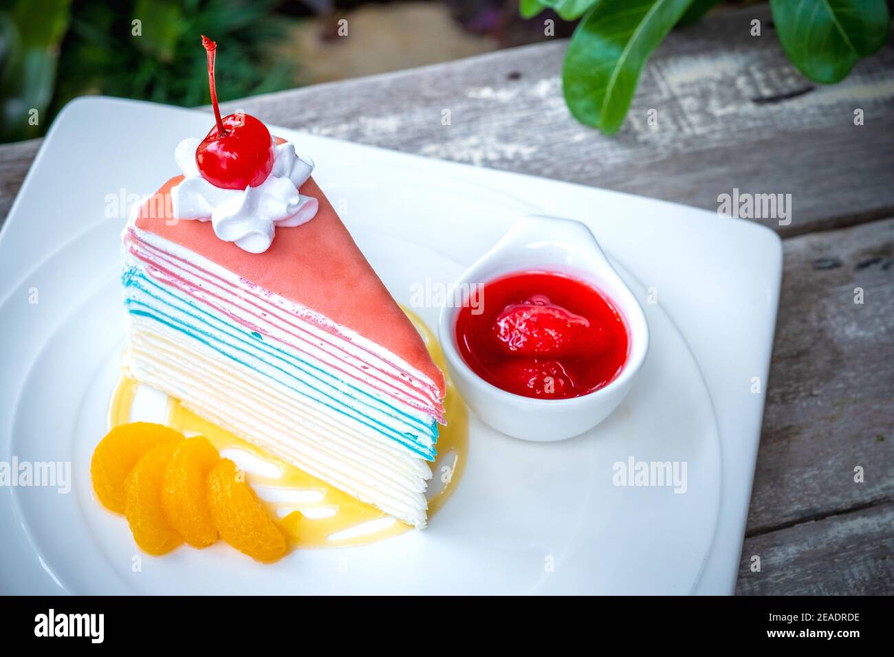 Gâteau à la crêpe arc-en-ciel et une cerise avec sauce fraise sur fond de table en bois. Banque D'Images