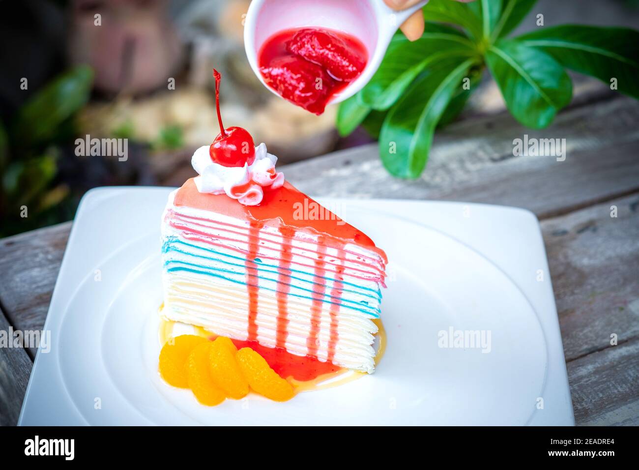 Gâteau à la crêpe arc-en-ciel et une cerise avec sauce aux fraises à la main sur le dessus, fond de table en bois. Banque D'Images