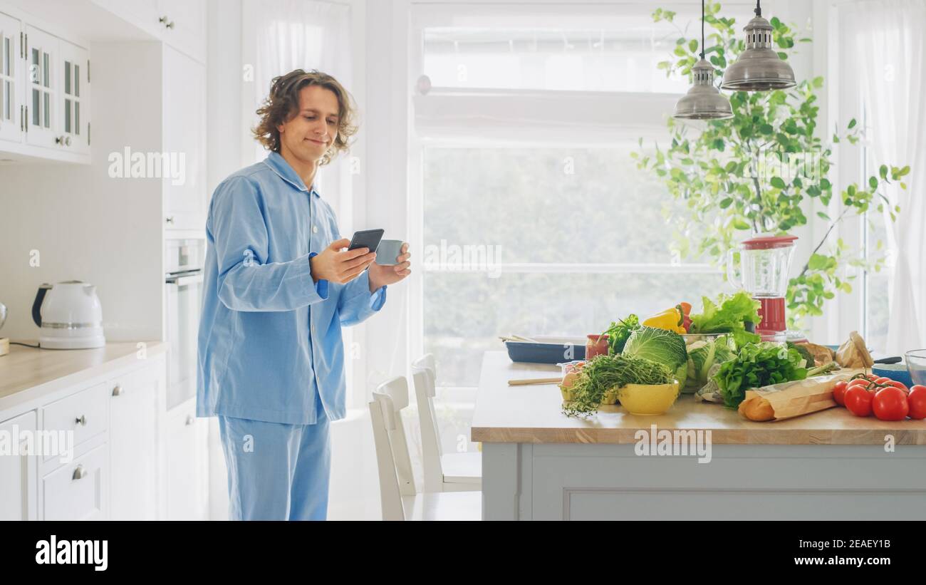 Happy Young Man with long Hair utilise un smartphone dans une cuisine tout en portant un pyjama bleu. Il fait défiler social Meadia et News Feed. Homme énergique Banque D'Images