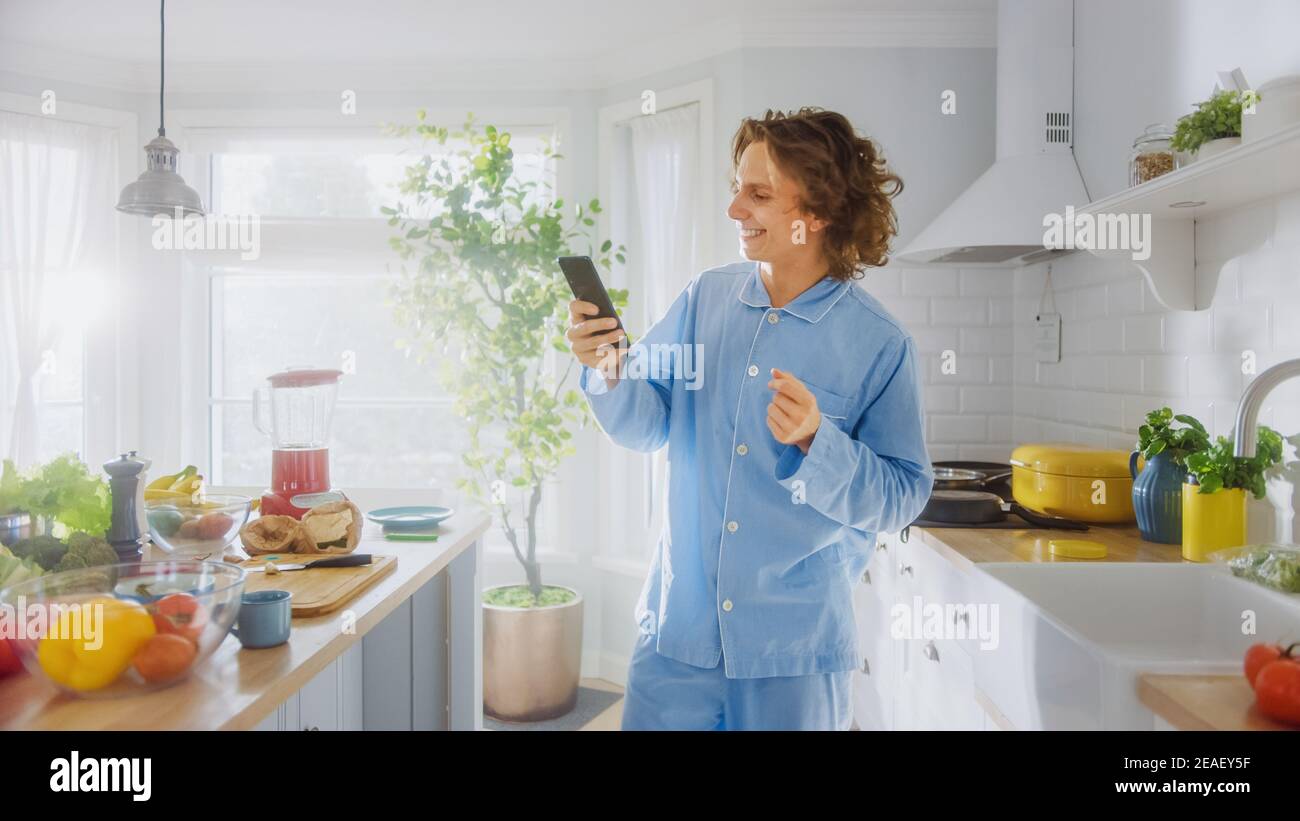 Happy Young Man with long Hair utilise un smartphone à la maison tout en portant un pyjama bleu. Energétique Man défilement des flux d'actualité et de contrôle des médias sociaux Banque D'Images