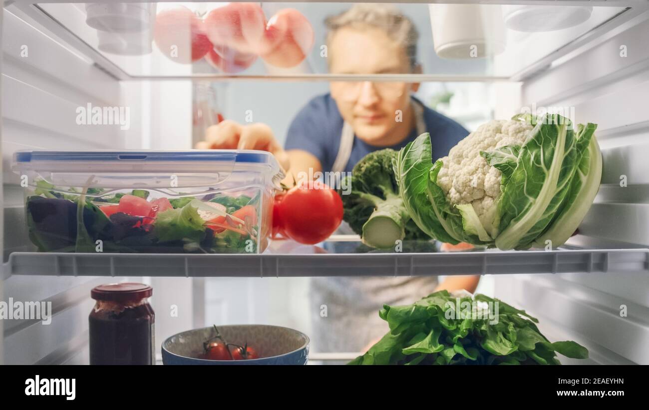 Beau jeune homme ouvre un réfrigérateur plein de nourriture biologique et saisit un bouquet de légumes pour une salade. Concept de régime alimentaire et de mode de vie sain. POV de Banque D'Images