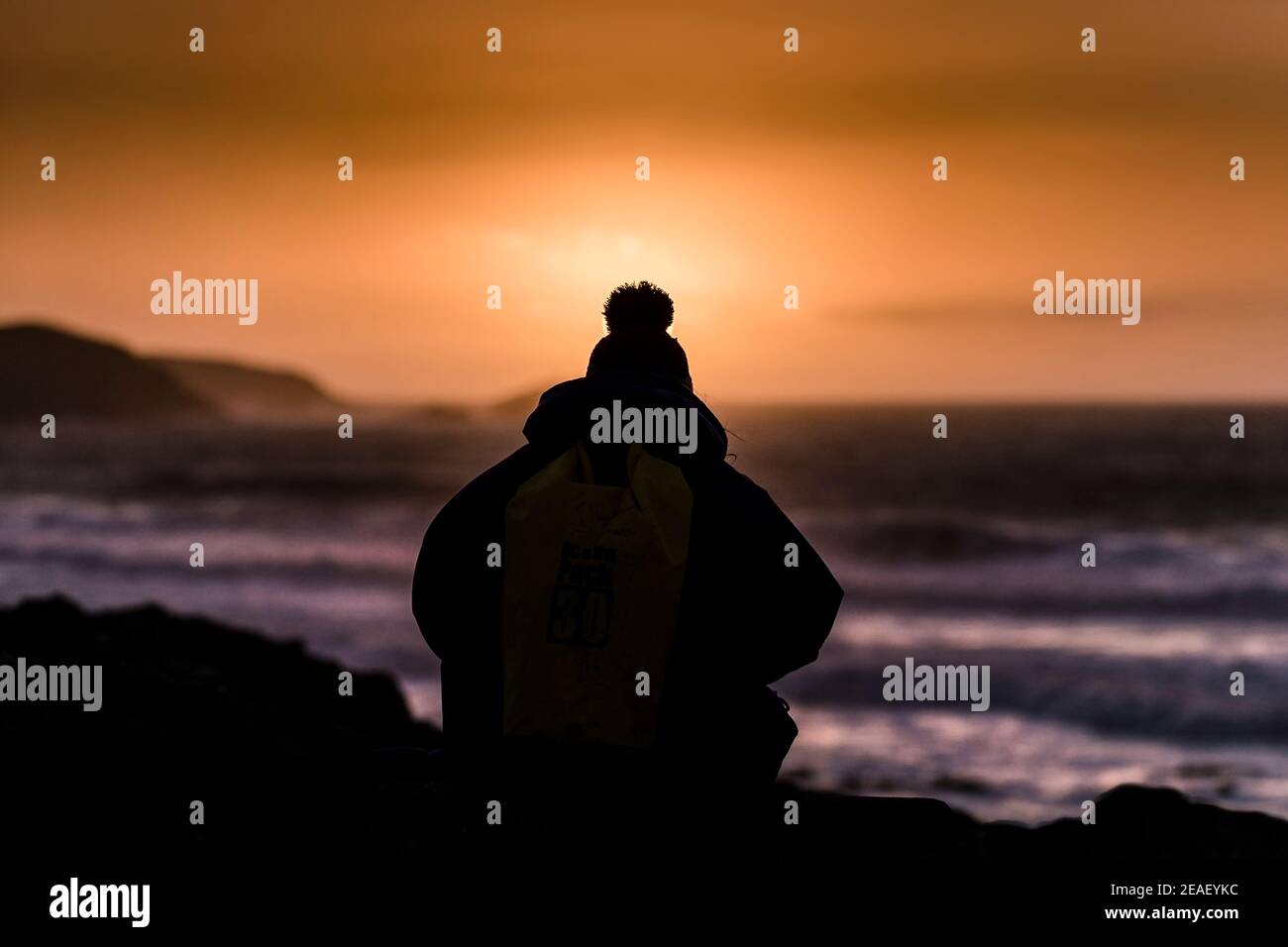 Une personne portant un chapeau de galet silhoueté par un coucher de soleil intense sur la côte de Newquay en Cornouailles. Banque D'Images