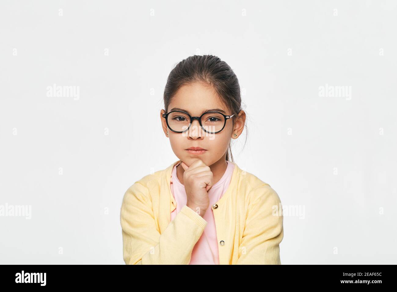 Portrait d'une petite fille de race mixte portant des lunettes rouges avec poing sous le menton, elle regarde sérieusement l'appareil photo Banque D'Images