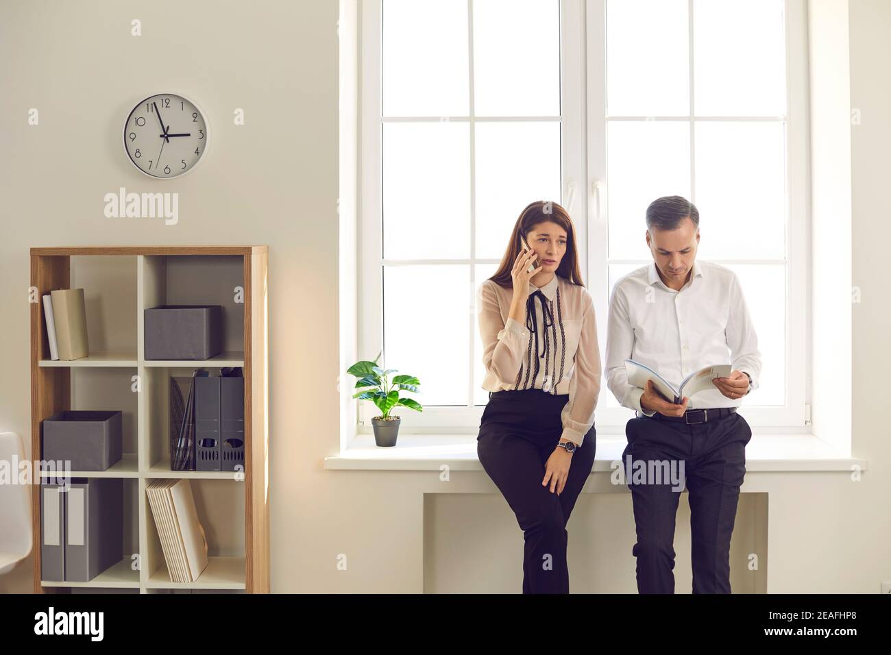 Homme regardant à travers les papiers et femme parlant au téléphone debout par la fenêtre pendant la pause au travail Banque D'Images