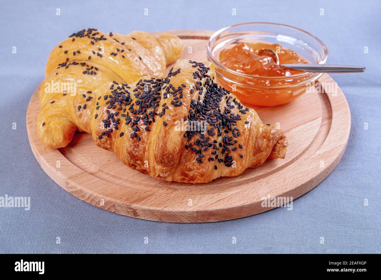 Deux croissants frais et un bol de confiture en verre sont sur une planche à découper ronde en bois ou en bois naturel sur une table avec une nappe en lin. Sélectif Banque D'Images