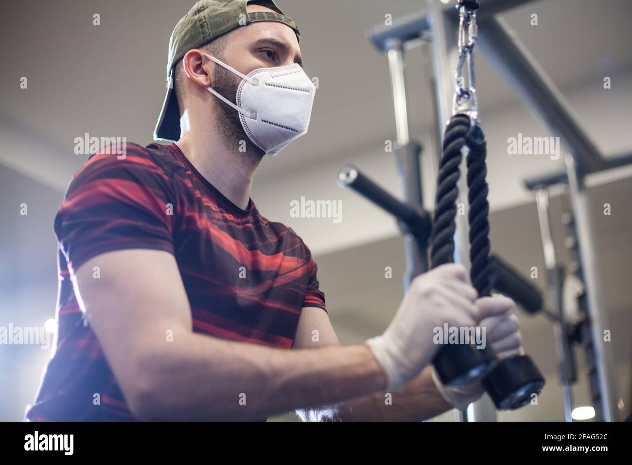 Jeune homme caucasien travaillant sur la machine de traction lat vers l'intérieur Rouvrez la salle de gym américaine, en portant des gants de protection en latex et un masque facial, activité et Maintien de la forme sous CO Banque D'Images