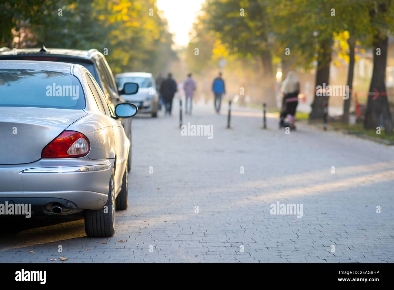 Voitures garées dans une rangée sur le côté de la rue de la ville le jour d'automne lumineux avec des personnes floues marchant sur la zone piétonne. Banque D'Images