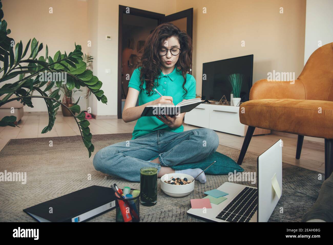 La fille caucasienne avec les cheveux bouclés a la leçon en ligne sur le sol à l'ordinateur portable tout en mangeant des céréales et du vert frais jus Banque D'Images