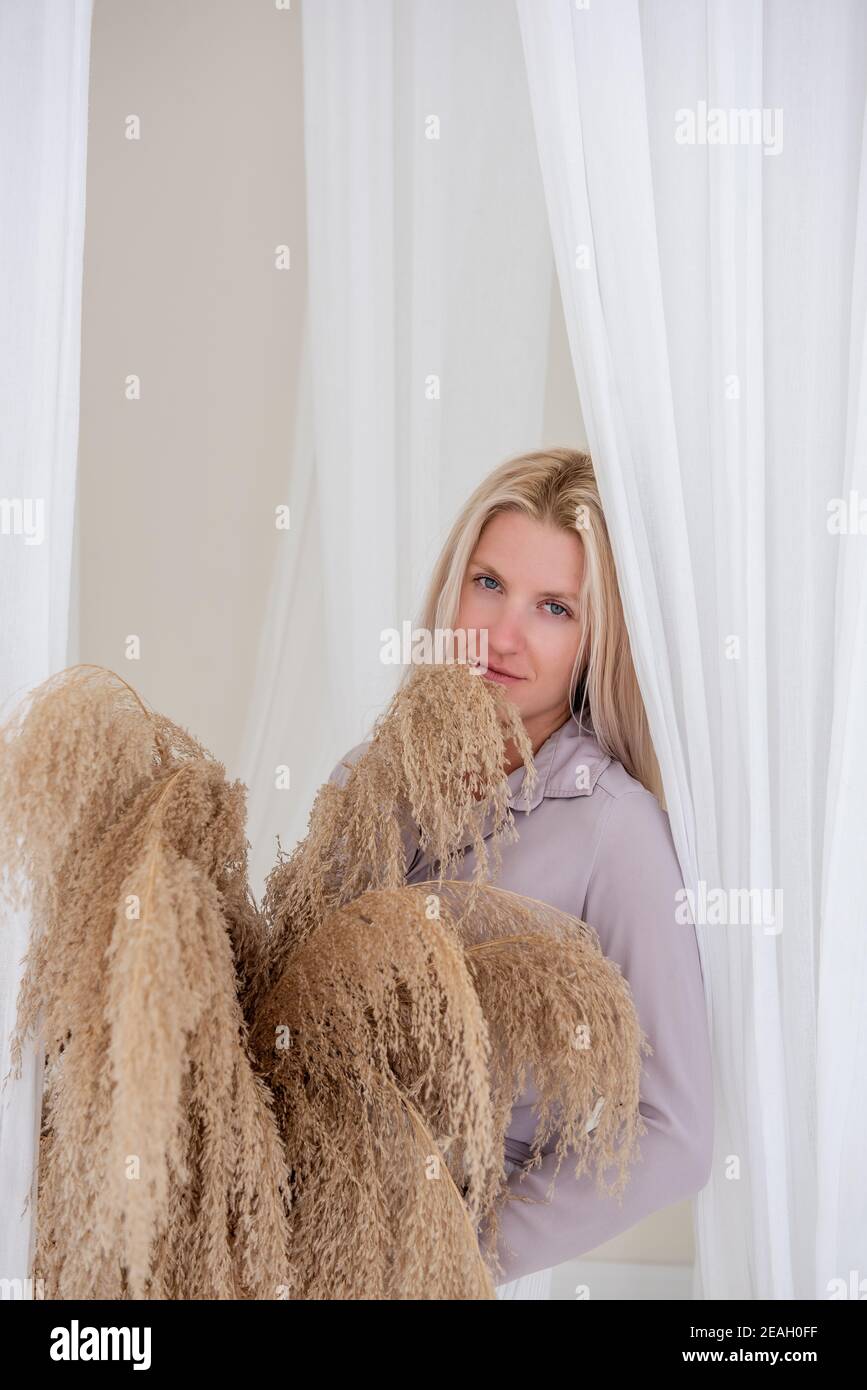 Jeune femme blonde dans une chemise lilas, robe blanche, tient dans ses mains panier de paille avec l'herbe de pampas parmi les aérées, développant des tissus de tulle. Minimali Banque D'Images