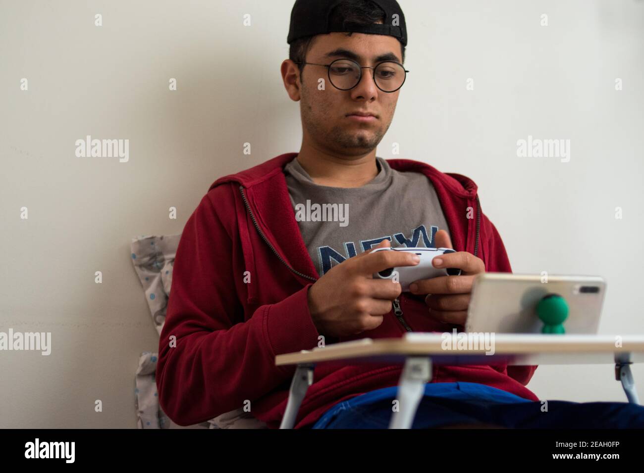 Jeune homme portant une veste rouge et des lunettes jouant à des jeux vidéo avec une manette blanche sur son smartphone à la maison Banque D'Images