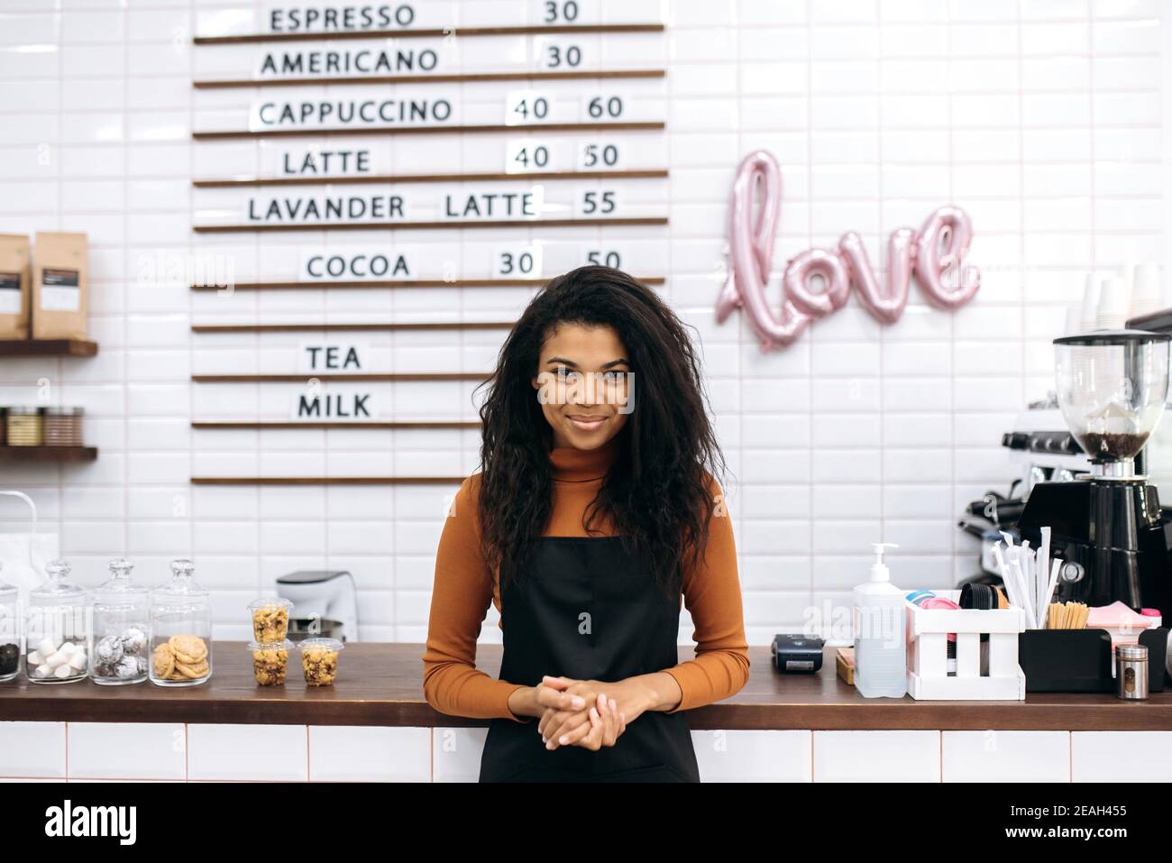 Concept de propriétaire d'entreprise de café. Jeune barista afro-américain ou serveuse en tablier noir souriant à l'appareil photo près du comptoir du café. Banque D'Images