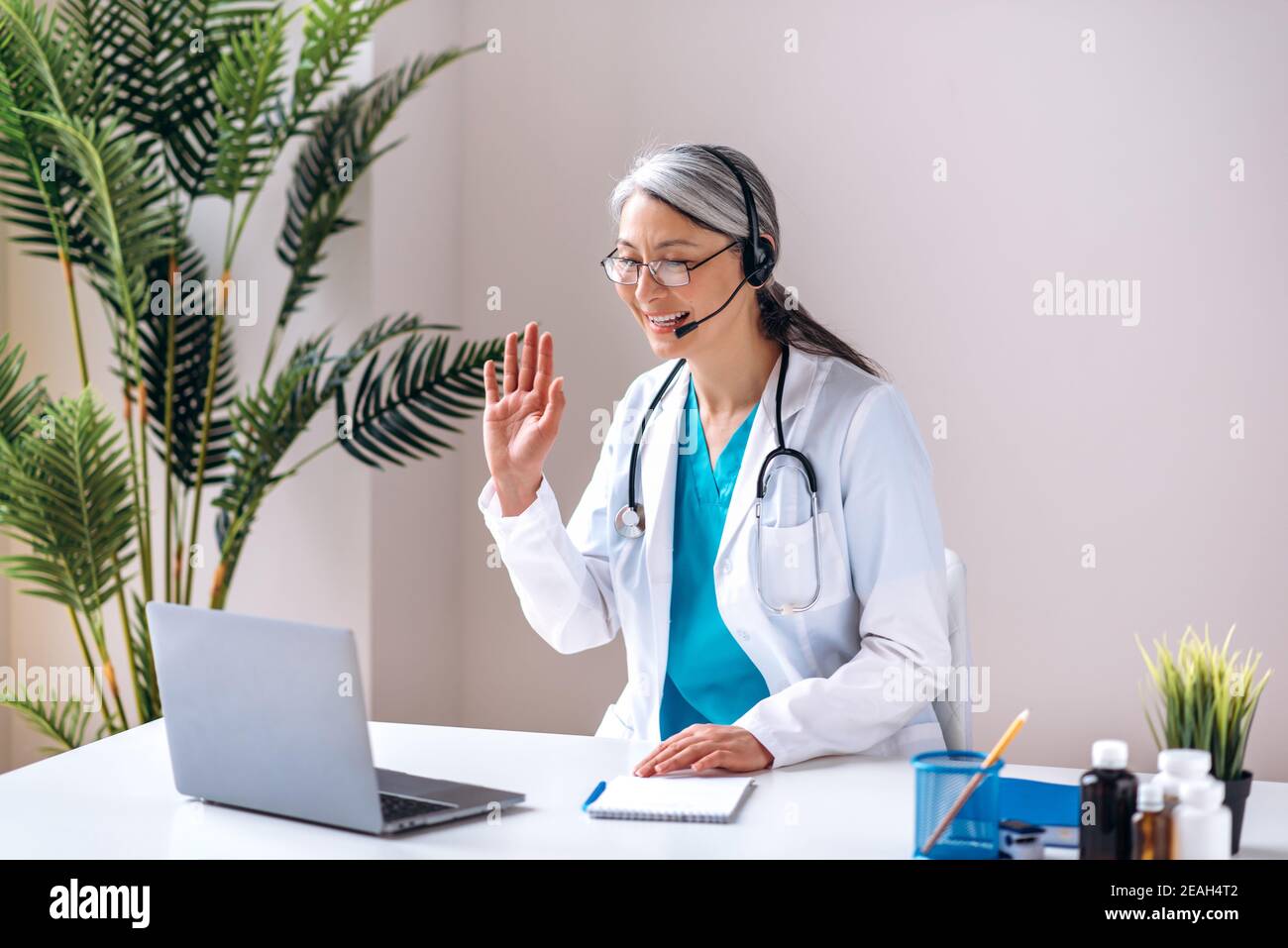 Une femme sympathique médecin dans un uniforme médical et des écouteurs consulte un patient en ligne, communique sur un appel vidéo à l'aide d'un ordinateur portable. Assistance médicale en ligne Banque D'Images