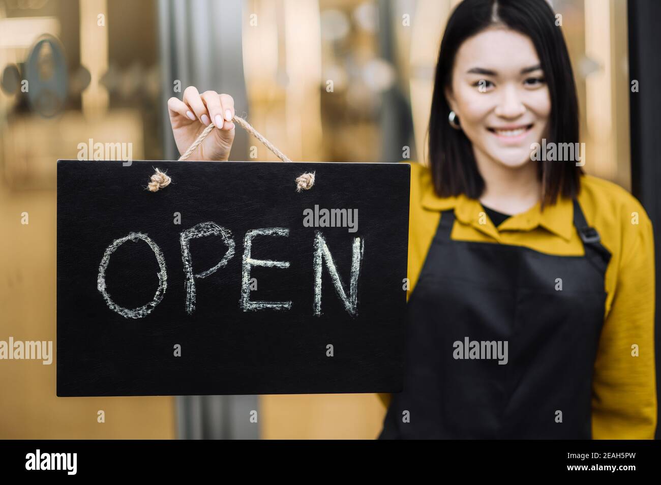 Nous sommes ouverts. Jeune femme, serveuse ou propriétaire d'un café ou d'un restaurant qui porte un tablier noir se tient à l'entrée avec une pancarte OUVERTE et souriante. Concept de petite entreprise Banque D'Images