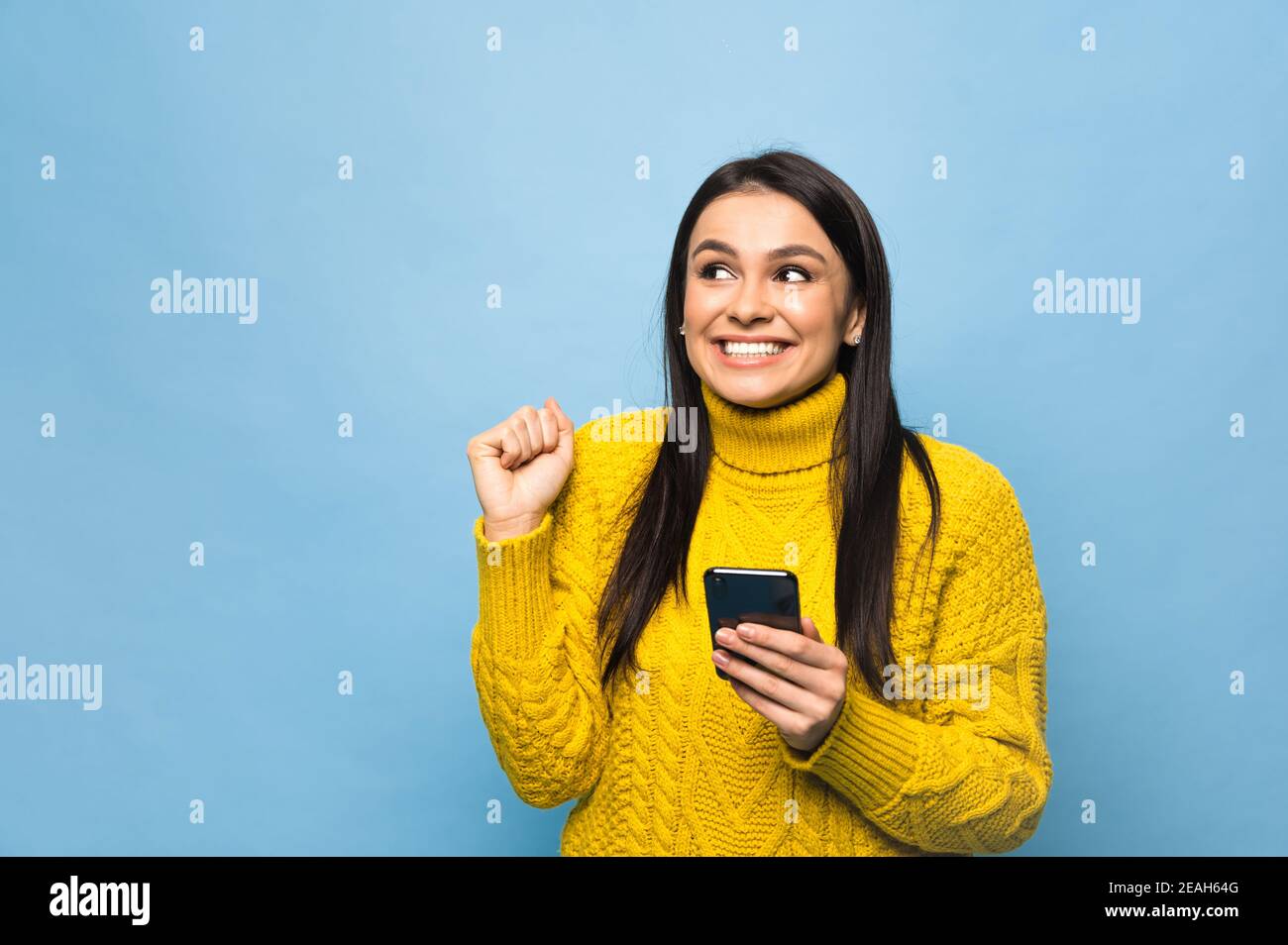 Portrait d'une jeune femme du caucase heureuse, tient le smartphone à portée de main, se réjouit d'une bonne nouvelle ou a reçu un message inattendu, se tient sur un fond bleu isolé Banque D'Images