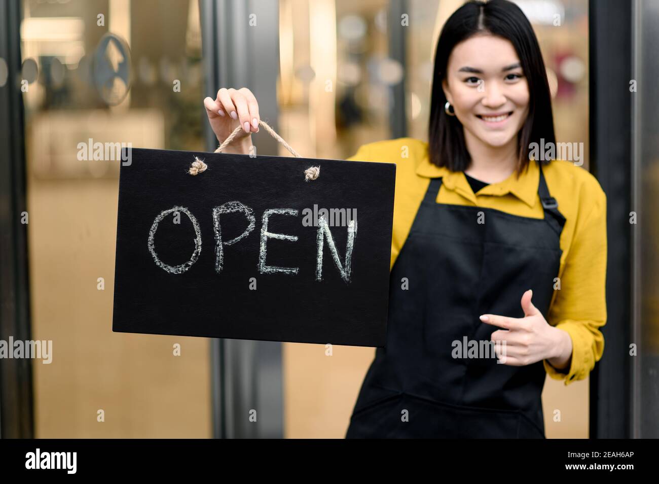 Bienvenue, nous sommes ouverts. Jeune femme, serveuse ou propriétaire d'un café ou d'un restaurant qui porte un tablier noir se tient à l'entrée avec une pancarte OUVERTE et souriante. Petite entreprise Banque D'Images
