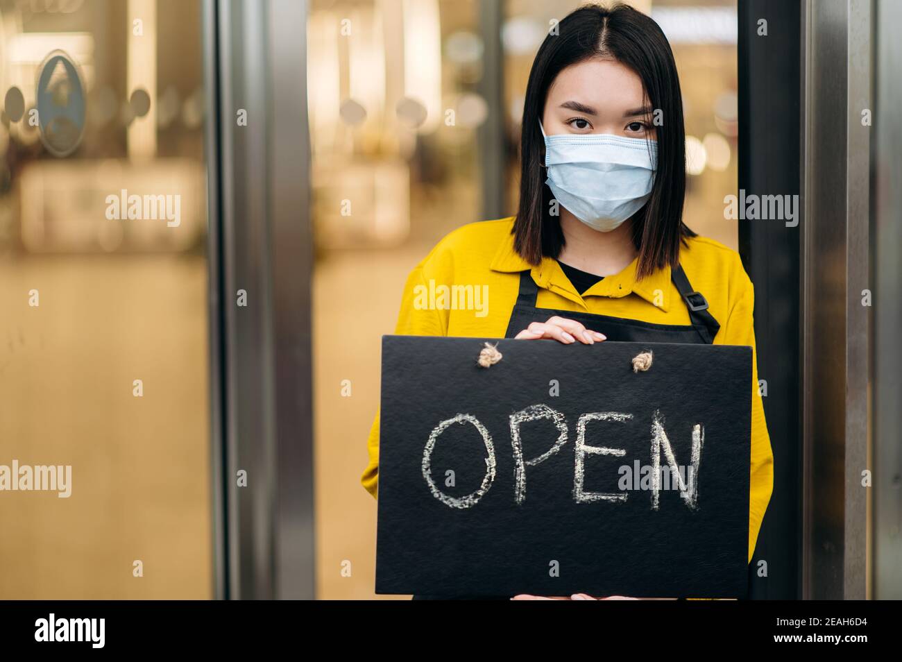 Portrait d'une serveuse heureuse debout à l'entrée du restaurant ou du café portant un masque médical sur son visage. Jeune femme asiatique montrant le signe ouvert dans sa petite boutique d'affaires et invite des clients Banque D'Images