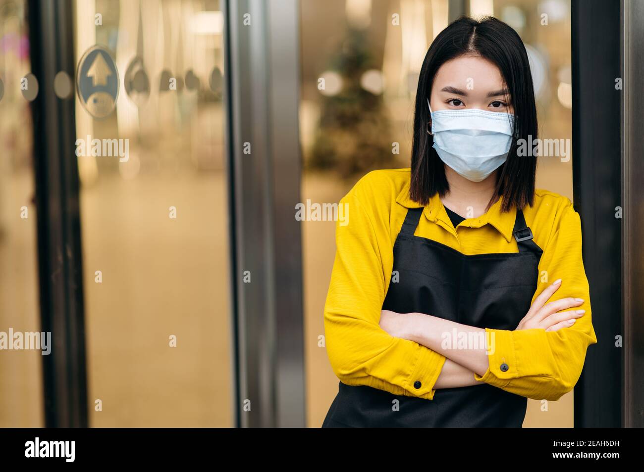 Une jeune femme asiatique serveuse ou propriétaire d'un restaurant, d'un café ou d'un magasin dans un masque protecteur et un tablier se tient à l'entrée. Petite entreprise dans le contexte de la pandémie du coronavirus Banque D'Images