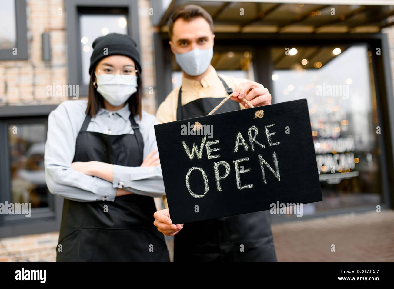 Panneau ouvert au premier plan. Deux serveurs défocused portant des masques protecteurs, homme caucasien et femme asiatique debout devant un restaurant, un café ou un bar et montrant le panneau OUVERT, bienvenue Banque D'Images