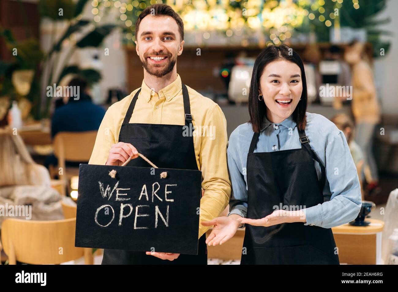 Portrait de deux serveurs sympathiques. Un jeune homme satisfait et une jolie jeune femme asiatique en tabliers noirs se tiennent à l'intérieur d'un restaurant, d'un café ou d'un bar, montrant le panneau OUVERT, avec un sourire agréable Banque D'Images