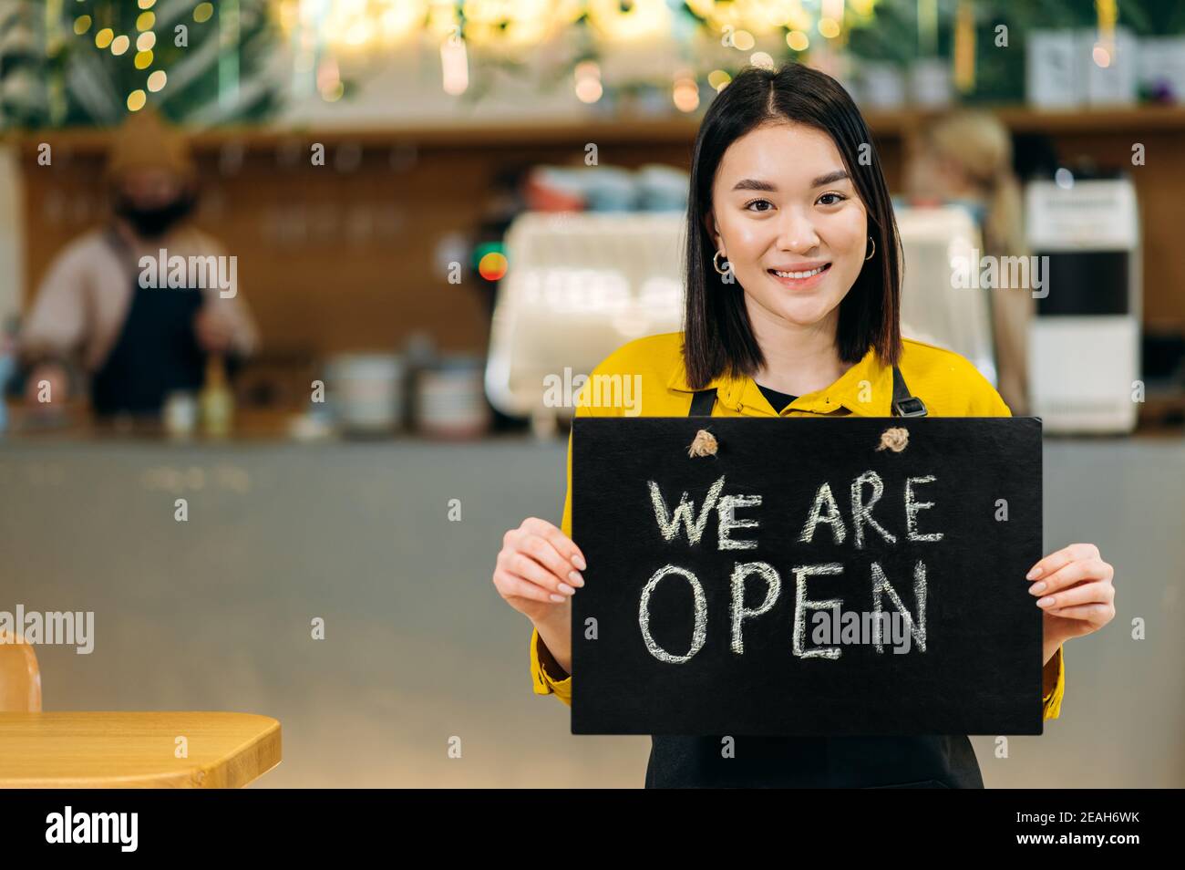 Nous sommes ouverts. Portrait d'une jolie serveuse asiatique heureuse debout à l'intérieur d'un restaurant ou d'un café portant un tablier noir tient UNE pancarte OUVERTE, et souriant sympathique Banque D'Images