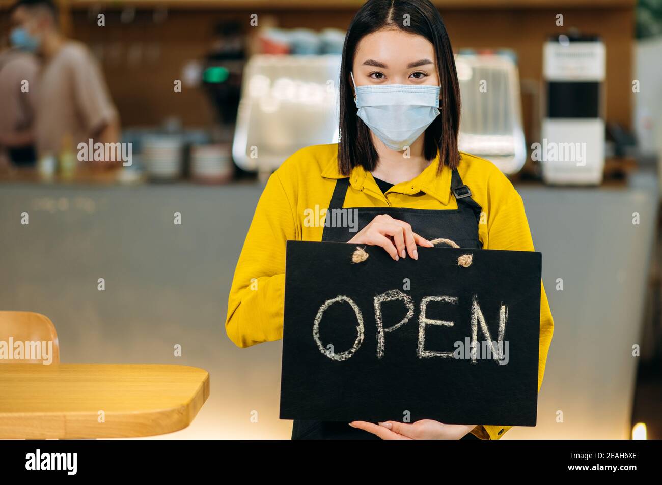 Panneau OUVERT. Portrait d'une jeune serveuse asiatique debout à l'intérieur d'un restaurant ou d'un café portant un masque médical de protection et un tablier noir et tient le panneau OUVERT. Prendre en charge le concept de petite entreprise Banque D'Images