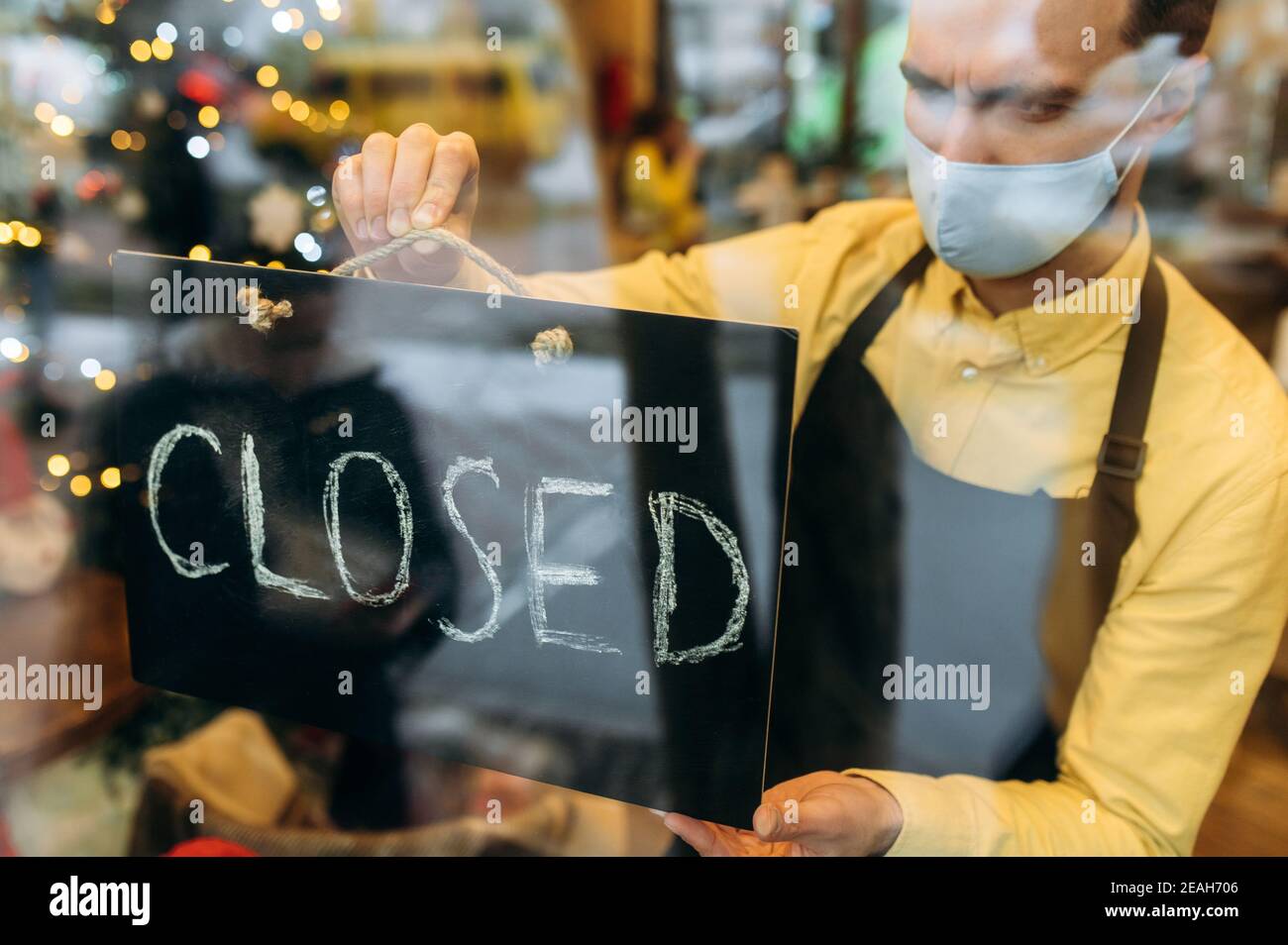 FERMÉ. Un serveur triste, un barista ou un propriétaire de petite entreprise portant un masque de protection médicale est accroché à un panneau FERMÉ à l'entrée d'un café, d'un restaurant ou d'un bar Banque D'Images