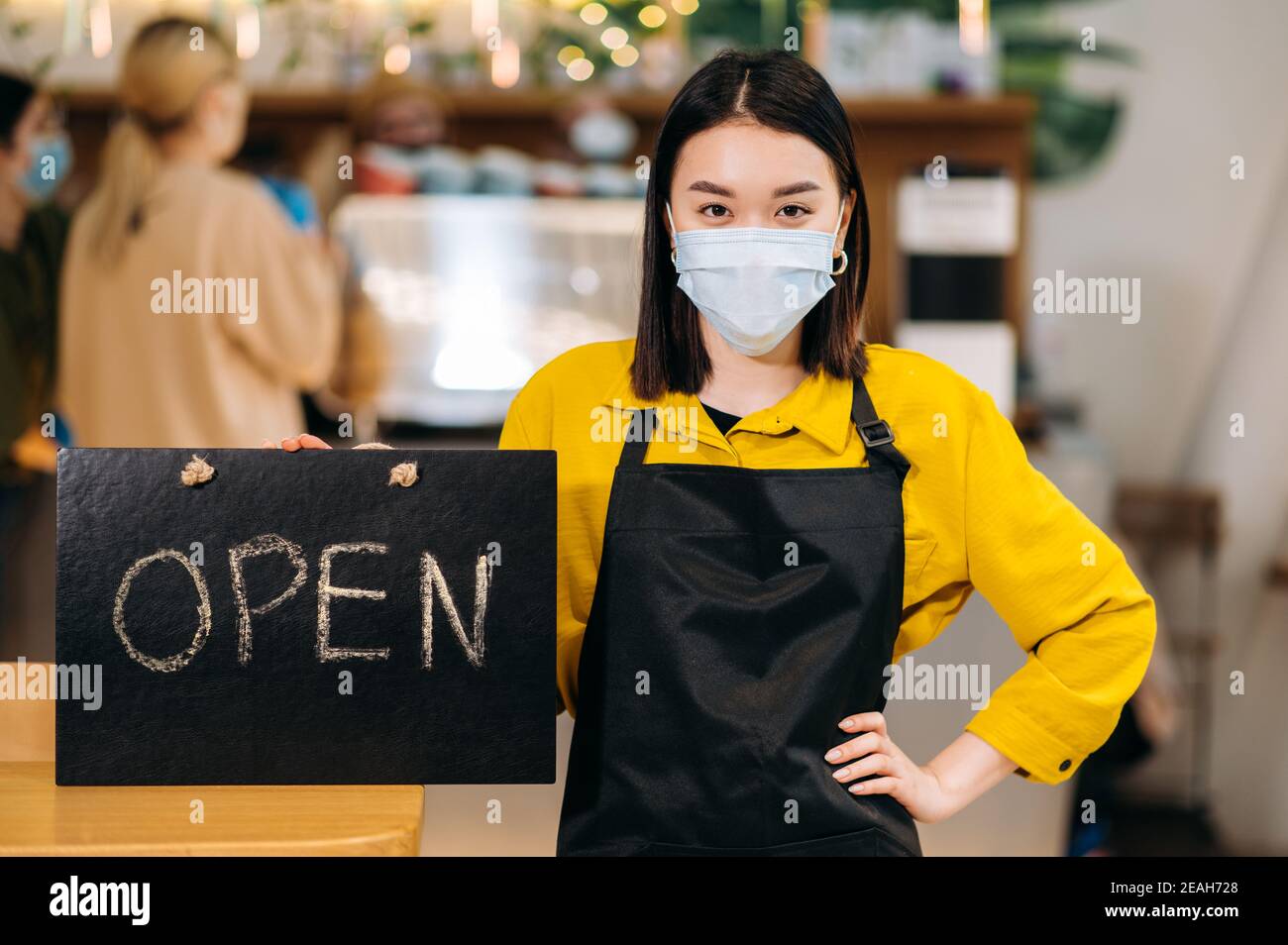 Bienvenue. Une jeune serveuse asiatique se tient à l'intérieur d'un restaurant ou d'un café portant un masque médical de protection et un tablier noir et tient le panneau OUVERT. Prendre en charge le concept de petite entreprise Banque D'Images