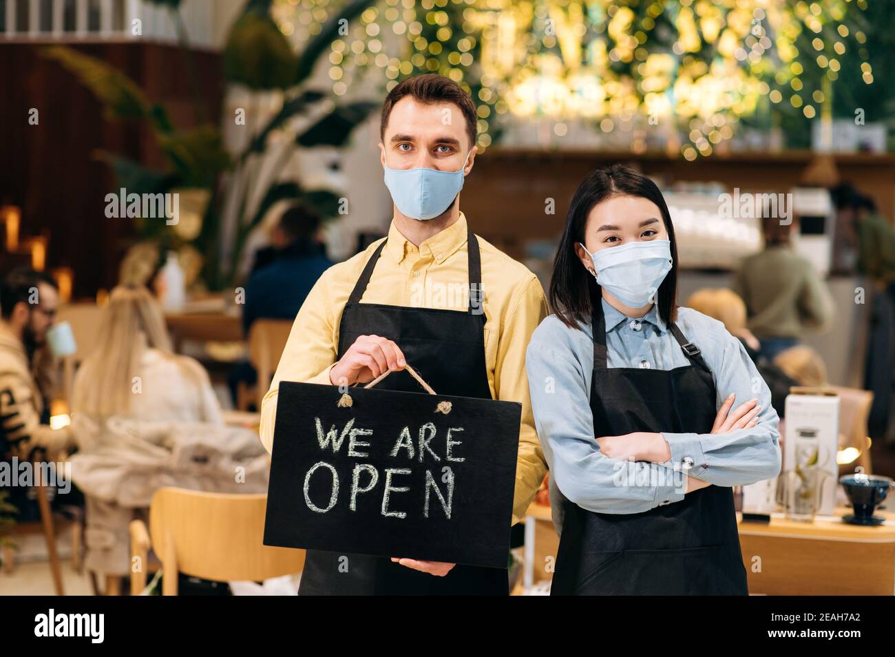 Bienvenue. Deux serveurs sympathiques portant des masques de protection. Un jeune homme satisfait et une jeune femme asiatique en tabliers noirs se tiennent à l'intérieur d'un restaurant, d'un café ou d'un bar et montrent le panneau OUVERT Banque D'Images