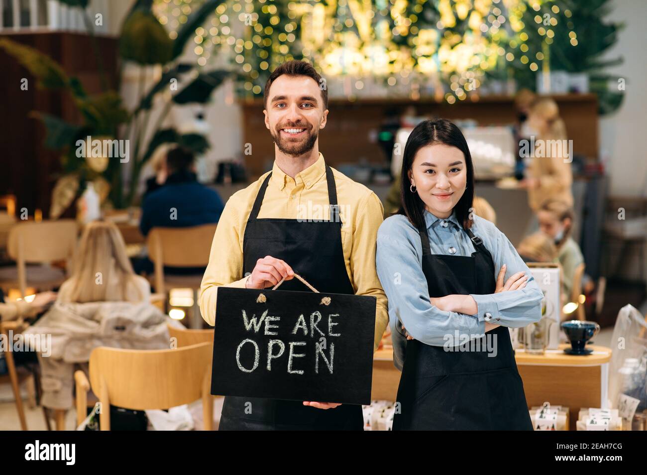 Portrait de deux serveurs sympathiques. Les employés multiraciaux satisfaits portent un uniforme dans un restaurant, un café ou un bar, montrant le panneau OUVERT, avec un sourire agréable Banque D'Images