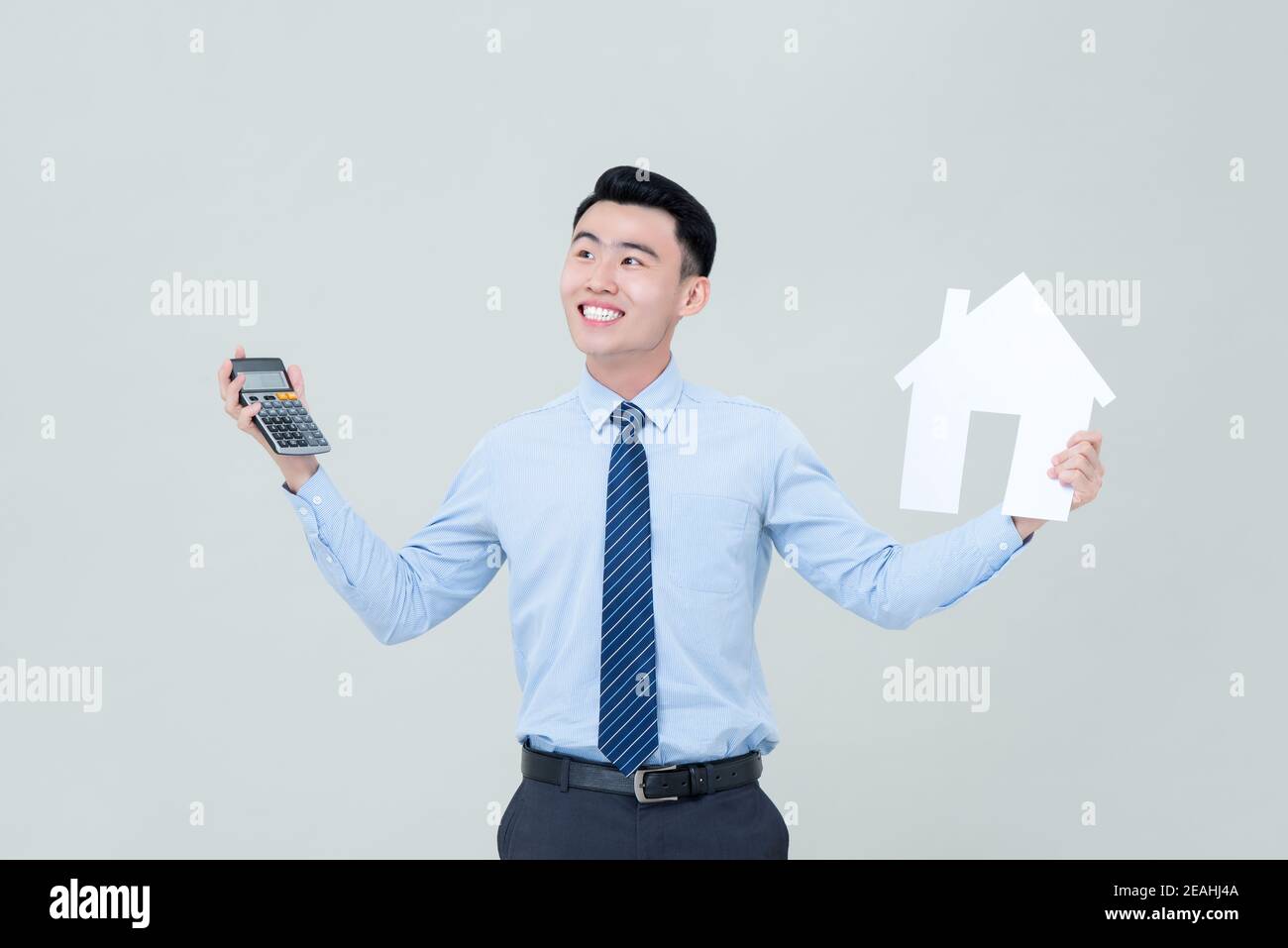 Jeune homme asiatique souriant agent immobilier tenant calculatrice et découpe de maison isolée sur fond gris clair Banque D'Images
