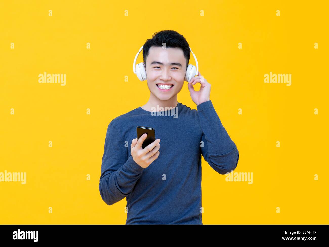 Un jeune homme asiatique souriant écoute de la musique en ligne depuis un smartphone isolé sur fond jaune Banque D'Images