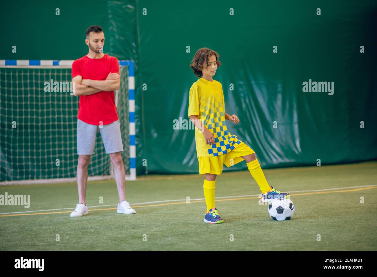 Mignon garçon en uniforme jaune jouant au football, son entraîneur le regardant Banque D'Images