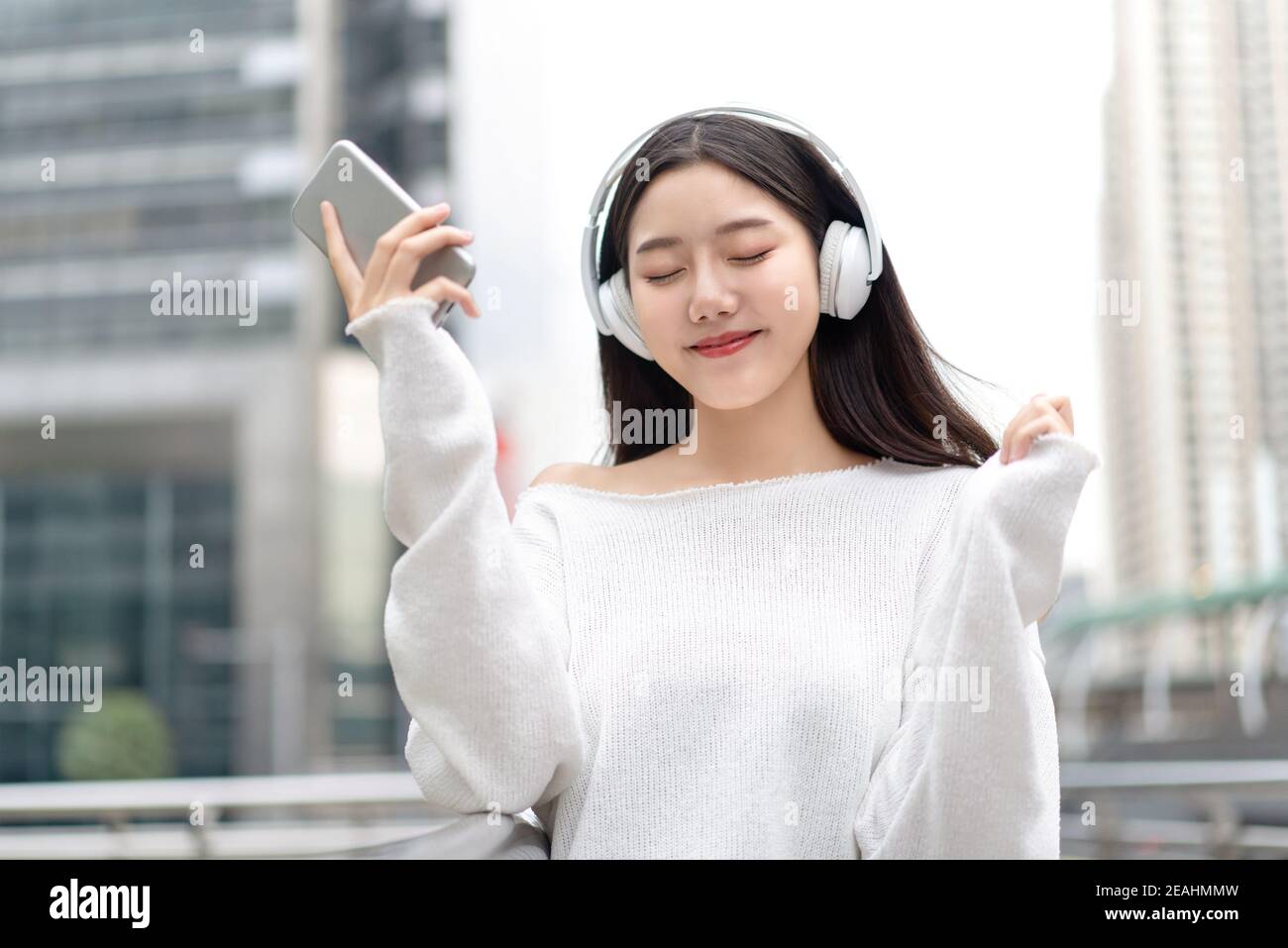 Bonne fille asiatique souriante portant un casque et écoutant de la musique en streaming avec des yeux fermés sur le fond du bâtiment de la ville Banque D'Images