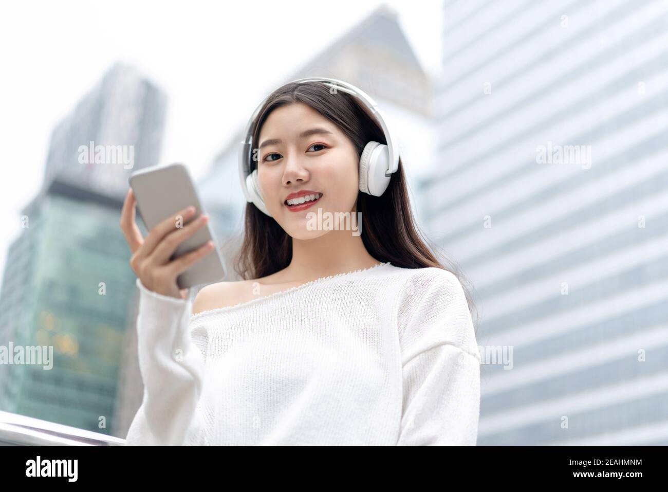 Bonne fille asiatique souriante, portant un casque pour écouter de la musique en streaming à l'extérieur, en arrière-plan du bâtiment de la ville Banque D'Images