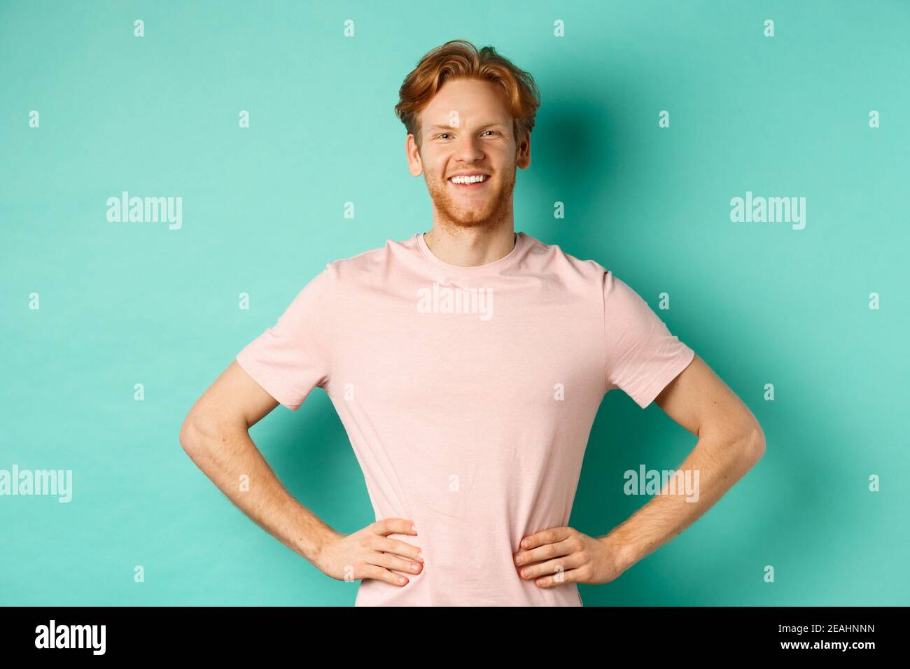 Jeune homme enthousiaste aux cheveux rouges, portant un t-shirt, se tenant heureux et fier des mains sur le houblon, debout sur fond turquoise Banque D'Images