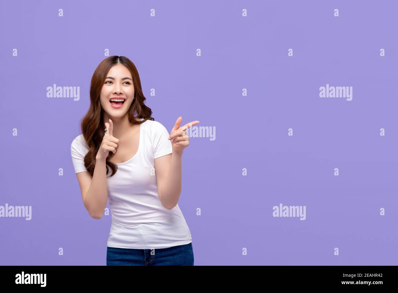 Jeune femme asiatique dans des vêtements décontractés avec grand sourire et bonne expression joyeuse pointant les mains vers l'espace vide isolé sur fond violet Banque D'Images