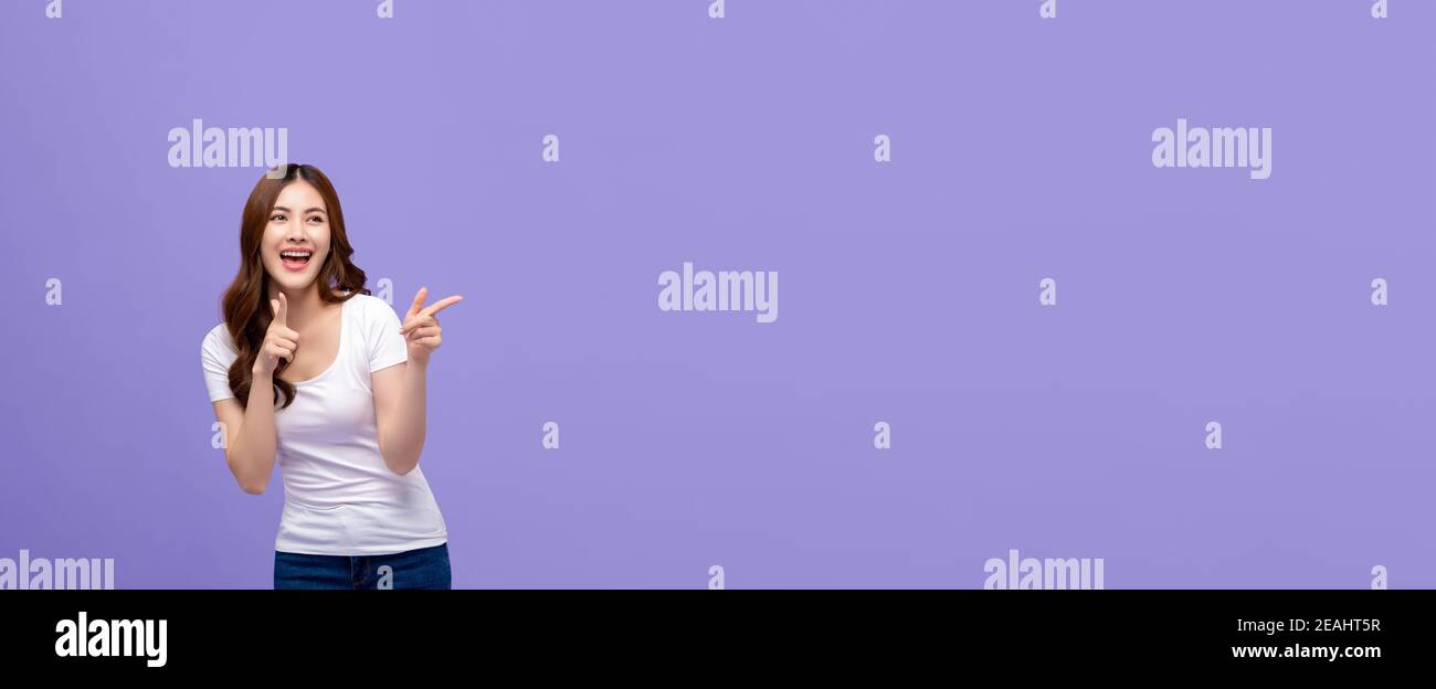 Jolie femme asiatique souriante dans un t-shirt blanc décontracté pointant de la main et en cherchant à copier l'espace de côté sur l'arrière-plan de la bannière violette Banque D'Images