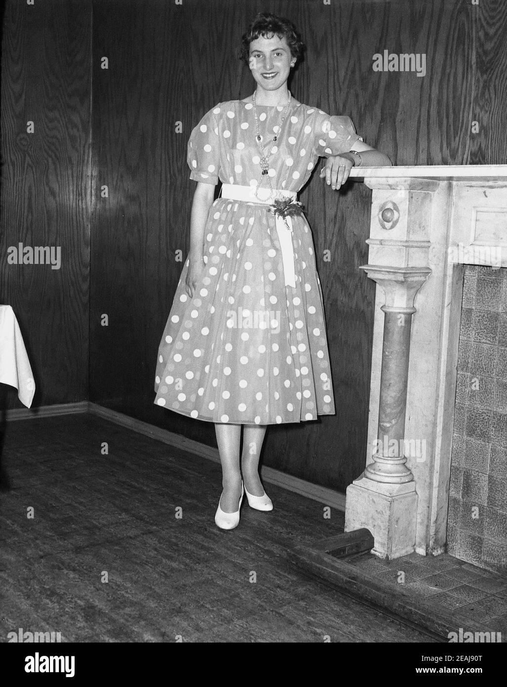 années 1950, historique, une jeune femme portant une robe à pois ou à pois debout dans une salle de réception à côté d'une cheminée. Elle célèbre son 21at anniversaire. Une telle robe de fête avec des points sur un fond avec une ceinture autour du milieu était à la mode à l'époque de l'oscillation rétro et de l'ère rockabiliy, l'un des premiers styles de rock et de musique de rouleau. Banque D'Images
