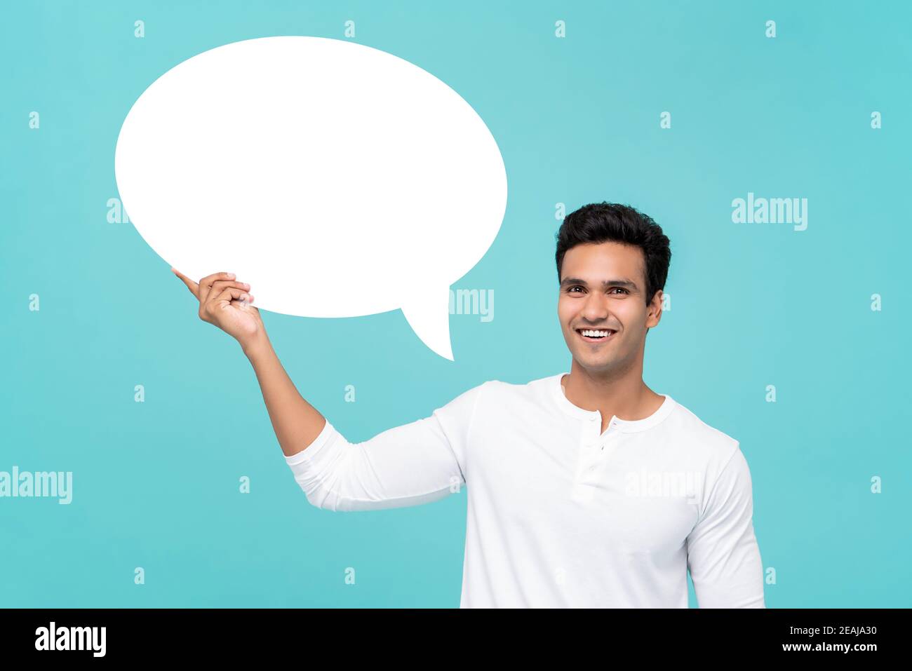 Beau homme indien souriant dans un t-shirt blanc tenant un discours blanc signe de bulle isolé sur fond bleu clair de studio Banque D'Images