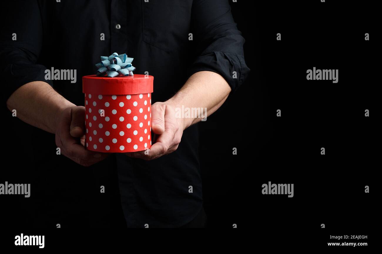 un homme adulte dans une chemise noire tient dans la main une boîte ronde rouge de pois avec un bleu arc Banque D'Images